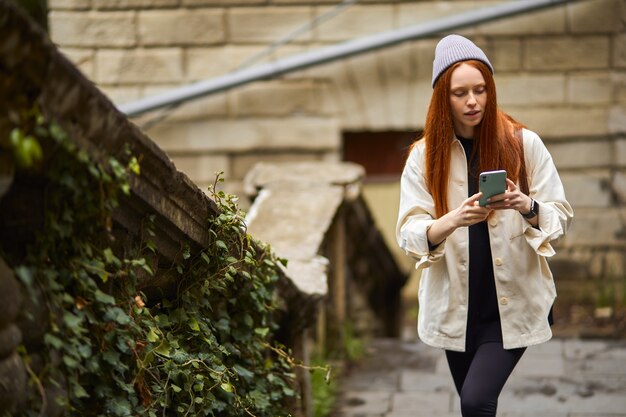 Femmina caucasica focalizzata usa lo smartphone mentre cammina, alla ricerca di un posto bellissimo tramite la mappa online sul telefono cellulare. Ritratto di signora dai capelli rossi in cappotto e cappello incentrato sulla ricerca