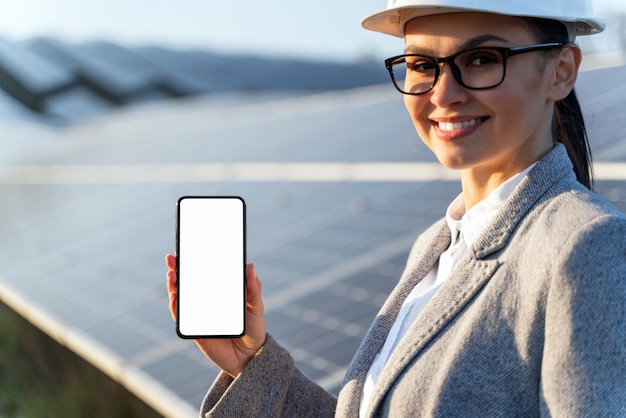 Femmina caucasica sorridente che mostra il suo telefono cellulare alla macchina fotografica davanti alle batterie di energia sul prato