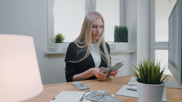 Femmina che conta soldi nel luogo di lavoro dell'ufficio. La bella giovane donna bionda in vestito che si siede nell'ufficio allo scrittorio di legno leggero con il computer che si concentra sul conteggio del grande mucchio di incassa le mani