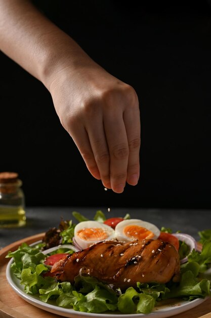 Femmina che fa un piatto di insalata aggiungendo una manciata di semi di sesamo bianchi su un petto di pollo alla griglia