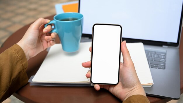 Femmina che tiene una tazza di caffè blu e uno smartphone con schermo bianco mockup alla sua scrivania portatile