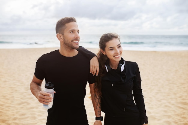 Femmina e maschio eccitati in abbigliamento sportivo con una bottiglia d'acqua che si divertono ad allenarsi sulla spiaggia in riva all'oceano