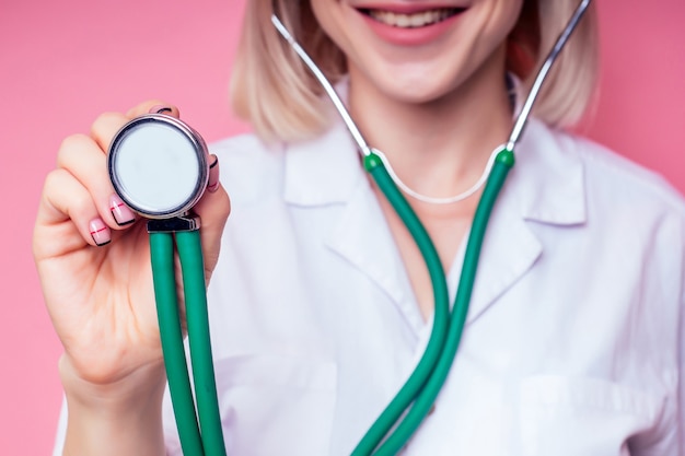 Femmina giovane e bella donna bionda medico veterinario che utilizza uno stetoscopio in un camice bianco su sfondo rosa in studio