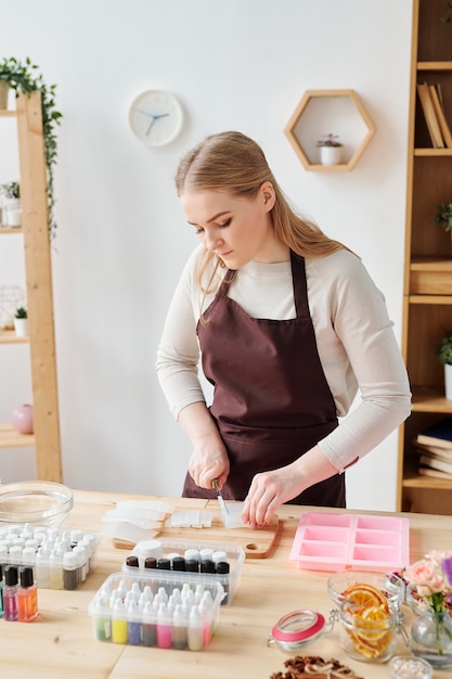 Femmina giovane produttore di sapone in grembiule marrone che taglia uno degli ingredienti sulla tavola di legno mentre si lavora in studio