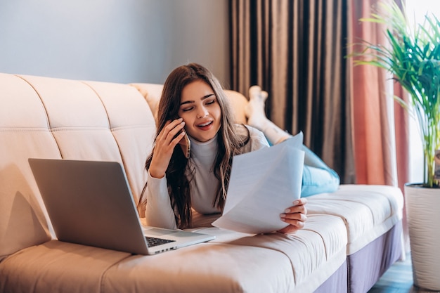 Femmina giovane studente freelance utilizzando laptop mentre era seduto a casa. Giovane donna che pone sul divano e lavora in linea sul computer portatile. Bella femmina parlando al telefono mentre studiava lavoro.