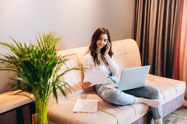 Femmina giovane studente freelance utilizzando laptop mentre era seduto a casa. Giovane donna seduta sul divano e lavora in linea sul computer portatile. Bella femmina parlando al telefono mentre studiava lavoro.