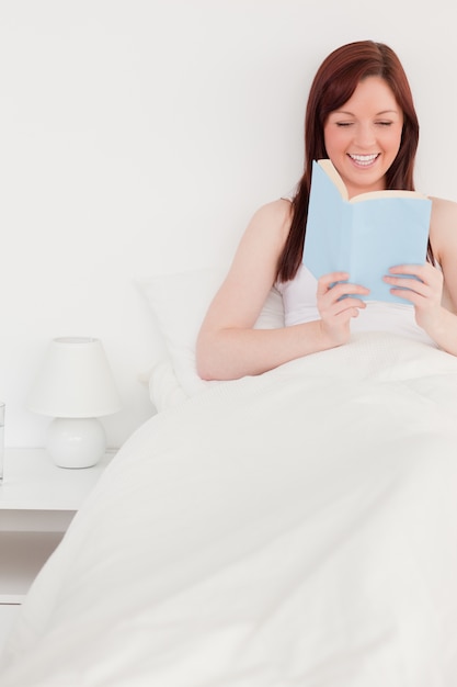 Femmina graziosa dai capelli rossi che legge un libro mentre sedendosi sul suo letto