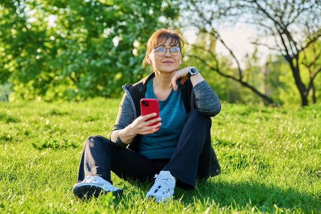 Femmina matura in cuffia con musica d'ascolto dello smartphone seduta sull'erba
