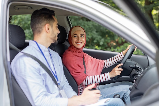 Femmina mediorientale millenaria sorridente in hijab al volante che guida un'auto che fa un esame di guida