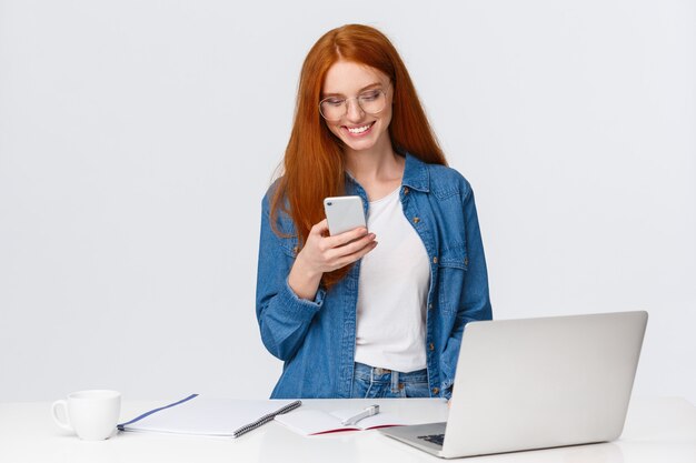 femmina rossa che lavora al progetto con lo smartphone, in piedi vicino al computer portatile