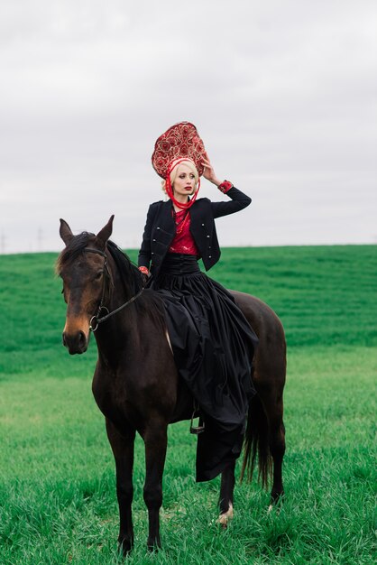 Femmina slava bionda in abito nero e copricapo kokoshnik sul campo con un cavallo nero al tramonto