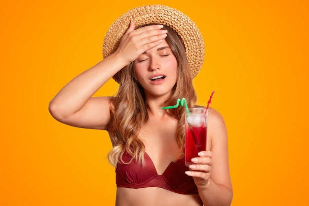 Femmina stanca in bikini e cappello estivo, tiene la mano sulla fronte, ha mal di testa dopo essere stata all'aperto durante le calde condizioni atmosferiche soffocanti