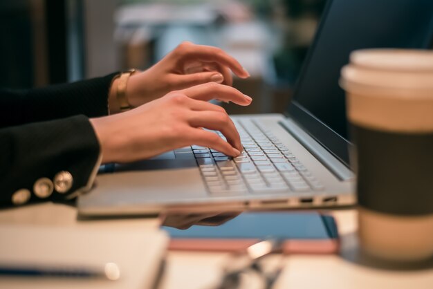 Femmina utilizzando il suo laptop in un bar. Vista laterale della giovane donna seduta a un tavolo con una tazza di caffè e telefono cellulare navigare in rete sul suo computer portatile.