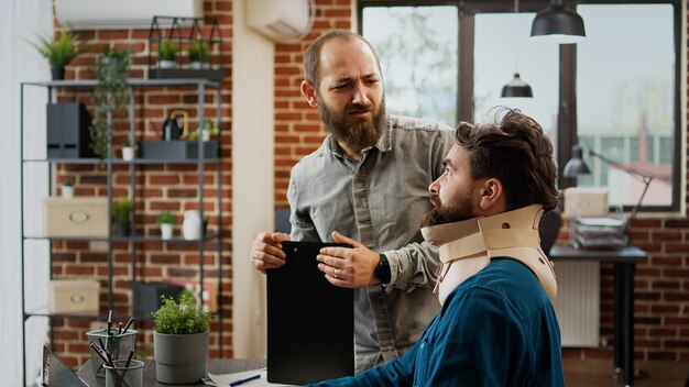 Ferito il dipendente dell'azienda con il collare cervicale dopo un incidente fisico, lavorando dolorante in ufficio. Uomo ferito che indossa un tutore medico in schiuma per il collo per curare la tensione e l'affaticamento alla schiena.