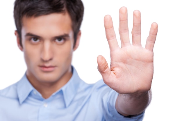 Fermare! Giovane serio in camicia blu che mostra il palmo della mano e guarda la telecamera mentre sta in piedi isolato su bianco