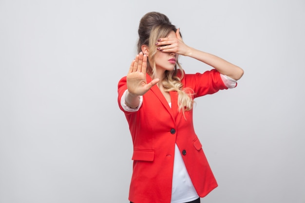 Fermati, non voglio vedere questo. Ritratto di bella donna d'affari con acconciatura e trucco in blazer rosso fantasia, in piedi coprendosi gli occhi con le mani di arresto. girato in studio, isolato su sfondo grigio.