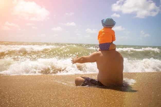 festa del papà. papà con un bambino sulle spalle in attesa di un'onda seduto sulla sabbia.
