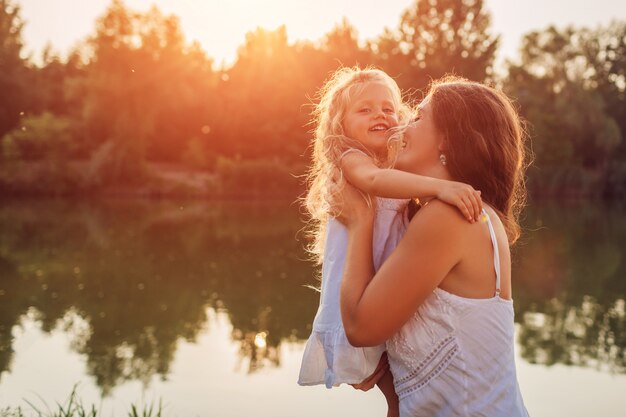 Festa della mamma. Donna che gioca divertendosi con la figlia dal fiume di estate al tramonto. Bambino e risata della tenuta della donna. Famiglia