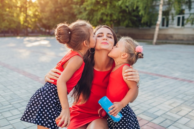 Festa della mamma. Figlie felici che baciano la loro madre dopo le lezioni all'aperto della scuola primaria con il presente. Abbraccio di famiglia. I bambini amano