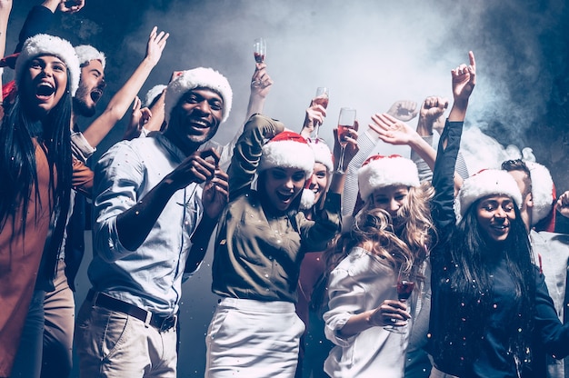 Festa di Capodanno con i migliori amici. Gruppo di bei giovani in cappelli di Babbo Natale che ballano e sembrano felici