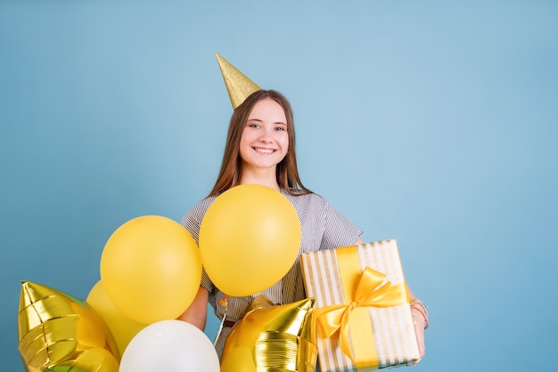 Festa di compleanno. Giovane donna in un cappello di compleanno che tiene palloncini e grande confezione regalo che celebra la festa di compleanno su sfondo blu con spazio di copia