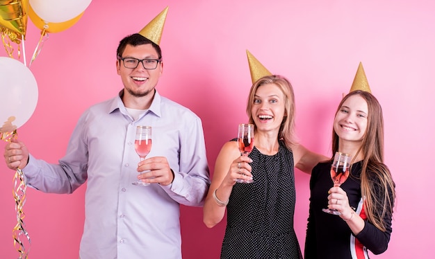 Festa di compleanno. Tre amici in cappelli di compleanno che bevono vino e celebrano la festa su sfondo rosa