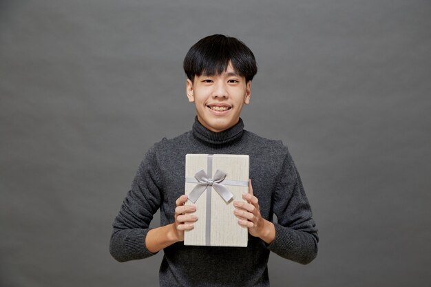 Festa di Natale e concetto di vacanze. Felice giovane uomo asiatico con scatola regalo studio shot, isolato su sfondo grigio