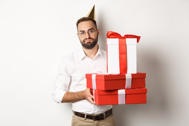 Feste e celebrazioni. Ragazzo dispiaciuto che tiene i regali di compleanno e che sembra deluso, non ama i regali, in piedi su sfondo bianco.