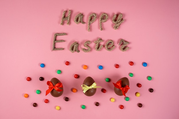Festeggia la Pasqua, la tipografia su corda e le uova di cioccolato tra i confetti colorati sullo sfondo rosa. Disegno di auguri.