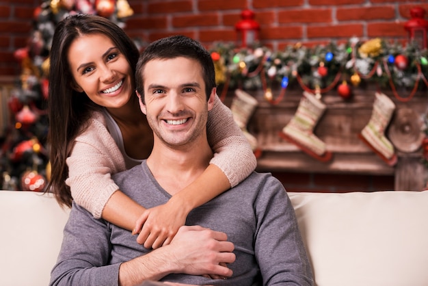 Festeggiamo il Natale insieme. Bella giovane coppia di innamorati che si legano l'un l'altro e sorridono mentre sono seduti sul divano con l'albero di Natale sullo sfondo