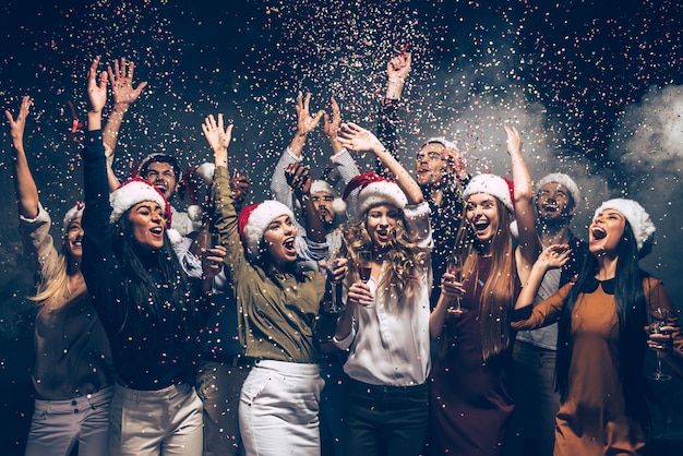 Festeggiamo il nuovo anno insieme. Gruppo di bei giovani in cappelli di Babbo Natale che lanciano coriandoli colorati e sembrano felici