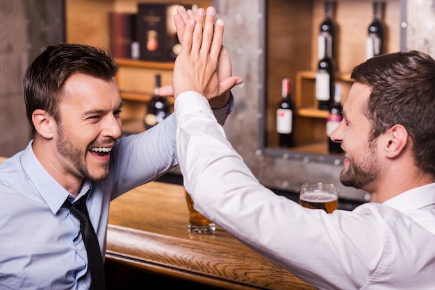 Festeggiamo insieme il successo. Due giovani allegri in camicia e cravatta che parlano tra loro e gesticolano mentre bevono birra al bancone del bar