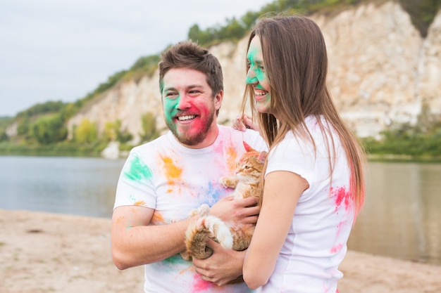 Festival holi, vacanze, turismo e concetto di natura - Coppia vestita con camicie bianche che tengono un gatto e ricoperte di polvere colorata.