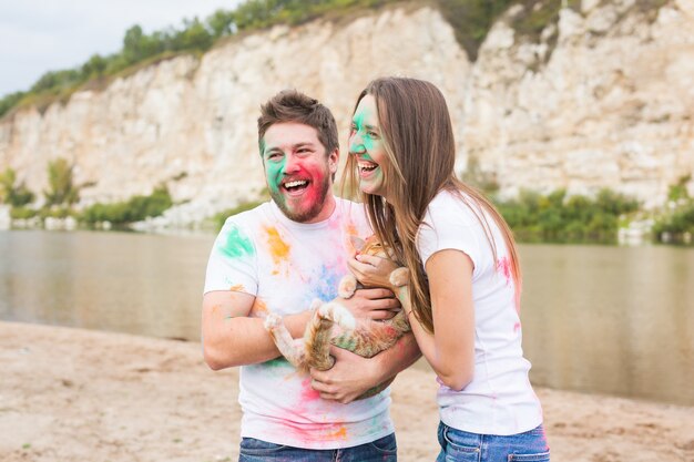 Festival holi, vacanze, turismo e concetto di natura - Coppia vestita con camicie bianche che tiene gatto e ricoperta di polvere colorata