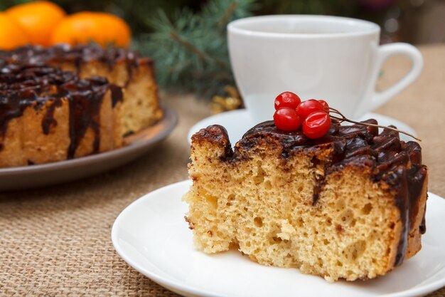 Fetta di torta al cioccolato decorata con grappolo di viburno, tazza di caffè, arance sul tavolo con ramo di abete e tela di sacco.