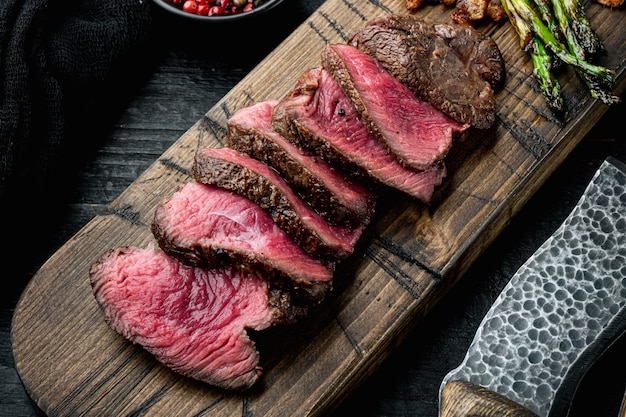 Fette di bistecca di carne marmorizzata alla griglia Filet Mignon set, con cipolla e asparagi, sul tagliere di legno, con coltello per carne e forchetta, sul tavolo di legno nero