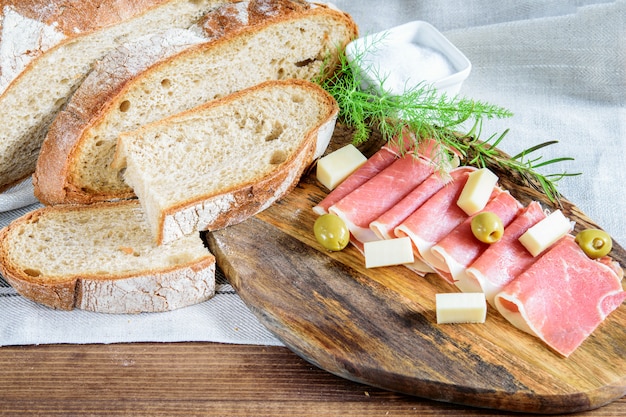 Fette di jamon, pane integrale, olive e formaggio sul tagliere di legno.