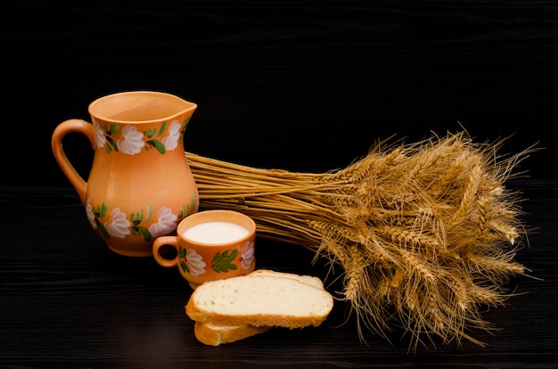 Fette di pane bianco, una tazza di latte e una brocca, un fascio di spighe di grano su un tavolo nero