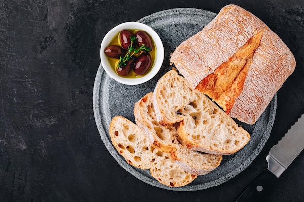 Fette di pane ciabatta italiano con olive su lastra di pietra su sfondo di cemento scuro vista dall'alto spazio copia