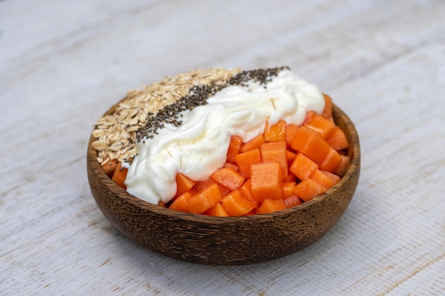 Fette di papaia dolce con fiocchi d'avena semi di chia e yogurt bianco in una ciotola di cocco su fondo di legno bianco da vicino