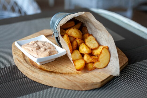 Fette di patate fritte rustiche in una busta di carta con salsa su un piatto di legno sulla terrazza all'aperto in un caffè.