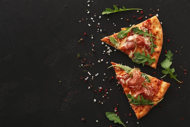 Fette di pizza con prosciutto e insalata di rucola con spezie su sfondo nero, copia spazio, vista dall'alto
