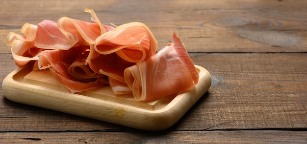 Fette sottili affettate di prosciutto sulla tavola marrone di legno, fondo di legno della tavola