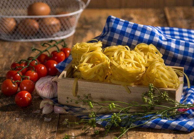 Fettuccine di pasta su tavola di legno con pomodorini, pepe, farina di frumento e uova in background.