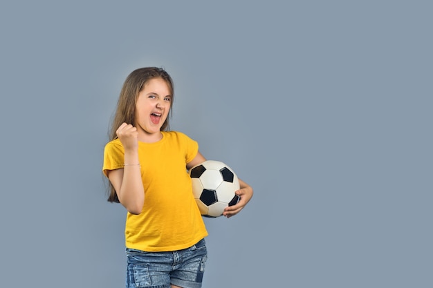 Ffan sport ragazza giocatore tenere il pallone da calcio celebrando, felice sorridente bambino in maglietta gialla su sfondo grigio, spazio di copia del testo libero