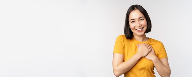 Fidanzata asiatica romantica che si tiene per mano sul cuore che sorride con cura e tenerezza in piedi in giallo ...