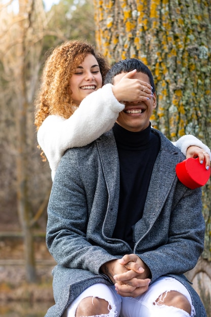 Fidanzata con proposta che copre gli occhi del fidanzato nel parco Giovane donna che tiene un regalo che copre gli occhi del suo fidanzato latino all'aperto