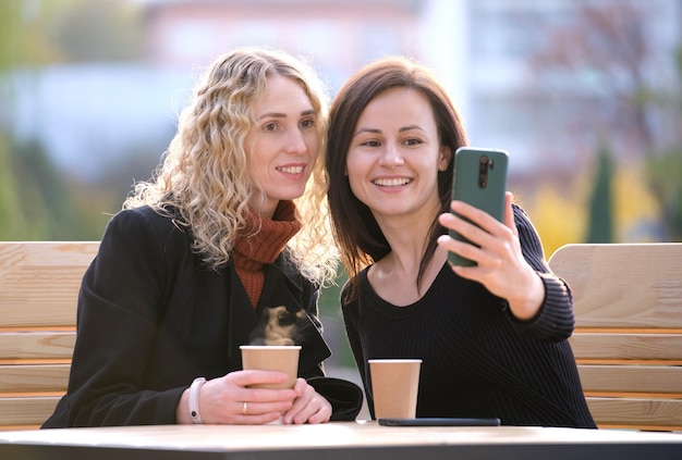 Fidanzate allegre che prendono selfie con il telefono che riposa al bar della strada della città Amici di sesso femminile che si godono il tempo insieme all'aperto in una calda giornata