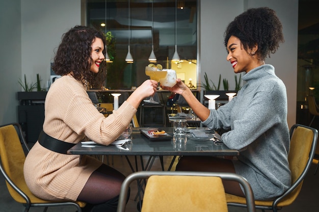 Fidanzate brindano sedute al tavolo brindando cocktail al ristorante - coppia di giovani amici che fanno una festa insieme