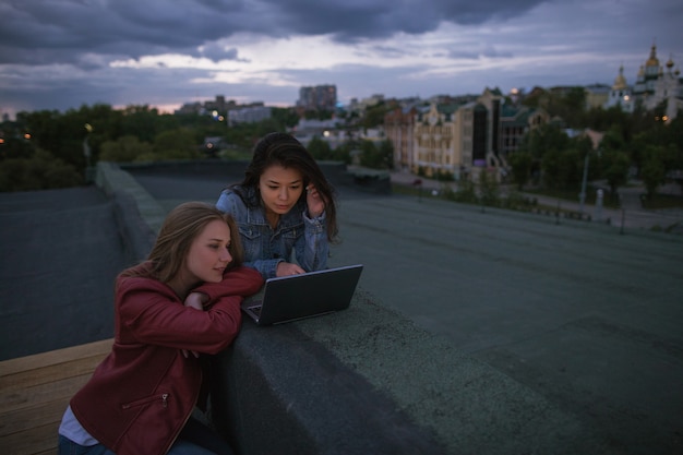 Fidanzate che guardano un film su un laptop seduto sul tetto contro un tempestoso cielo serale. Le ragazze comunicano in un social network, fanno acquisti online, lavorano da remoto